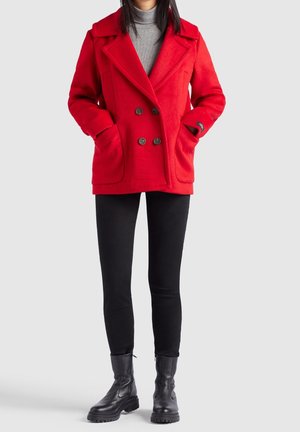 Kvinde iført en knaldrød dobbeltradet frakke, grå rullekrave, sorte bukser og sorte snørestøvler stående mod en ensfarvet baggrund.