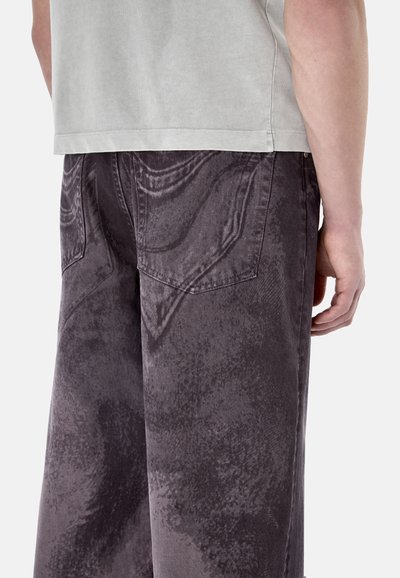 Person, die weite, dunkelgraue Jeans mit Acid-Waschung und ein hellgraues Kurzarmhemd trägt, ab Taille bis Mitte Oberschenkel, Rückansicht.