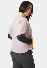 Sudadera de forro polar en rosa claro con cuello amarillo y mangas negras. Presenta un bolsillo frontal y un dobladillo curvado. Tejido suave y texturizado.