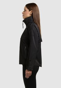 Femme aux longs cheveux bruns portant une veste noire à col montant avec poches zippées, debout de profil sur un fond clair.