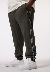 Donkergroene sweatpants met een slim fit, zijritszak en grijze strepen aan de zijkanten, gecombineerd met witte sneakers.
