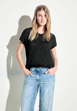 Schwarzes Kurzarmoberteil mit strukturiertem Auschnittmuster, kombiniert mit hellblauen Jeans. Der Hintergrund ist schlicht weiß.