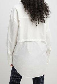 Chemise blanche surdimensionnée avec un panneau arrière boutonné, des manches longues et un ourlet arrondi. Texture lisse, associée à un pantalon à rayures sombres.