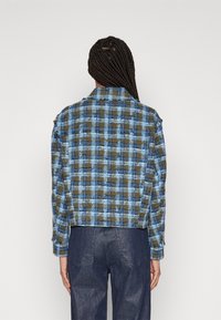 Kariertes Jackett in Grün und Blau, mit Kragen, langen Ärmeln und einem strukturierten Stoffmuster. Kombiniert mit dunklen Denim-Hosen.