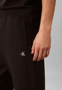 Parem käsi mustade pükste kõrval, mille reiel on valge tikitud „cK“ logo, halli taustaga.