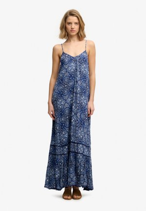 Jeune femme portant une maxi robe sans manches à motifs bleus avec de fines bretelles et des sandales marron à bout ouvert, debout devant un fond blanc.