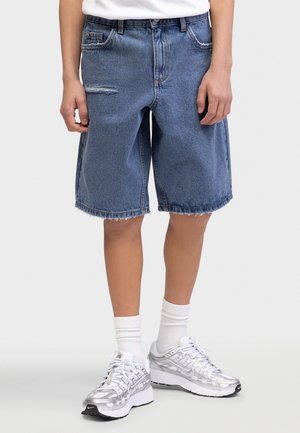 Person iført blå denim-knælange shorts med frynsete kanter, hvide sokker og sølv- og hvide sneakers, stående på ensfarvet baggrund.