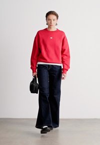 Sweat-shirt rouge, jeans foncés évasés, sac à main noir. Texture lisse, détail de logo sur la poitrine, design décontracté, debout contre un fond neutre.