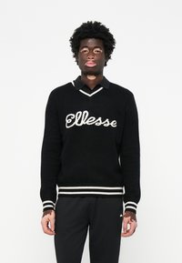 Sort strikket sweater med hvide striber ved �ærmer og kant, prydet med et fremtrædende "Ellesse" logo over brystet og en V-halsudskæring.