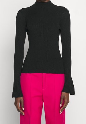 Femme portant un pull noir côtelé à col roulé ajusté et un pantalon taille haute large couleur rose vif, debout les bras détendus.