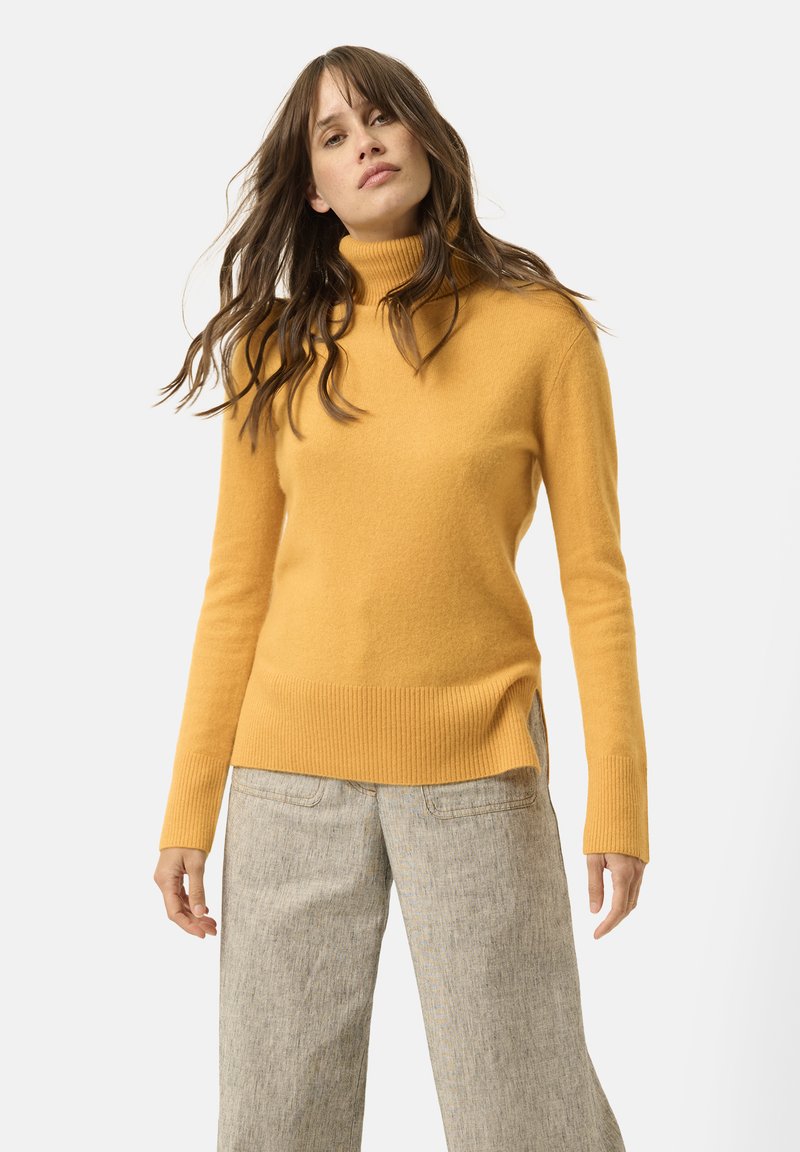 Maglione a collo alto giallo senape realizzato in materiale morbido; presenta orli e polsini a coste; abbinato a pantaloni ampi grigi chiari.