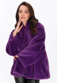 Femeie cu păr brunet, îmbrăcată într-o haină de blană purpurie pufoasă și pantaloni negri, atingându-și fața cu o mână, pe un fundal alb.