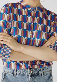 Blusa colorata e stampata con design geometrici blu, bianchi e marroni, colletto rialzato e chiusura a bottone in tessuto leggero. Parte inferiore in denim visibile.