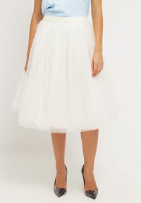 Falda de tul blanca con un diseño acampanado y cinturilla, que presenta una textura translúcida que proporciona un efecto de capas. Combinada con zapatos de tacón altos grises.