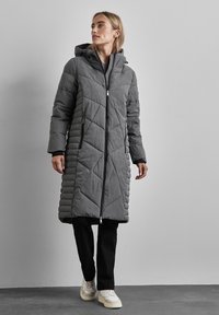 Manteau long gris matelassé avec capuche, présentant des coutures diagonales, fermeture zippée à l'avant et deux poches latérales. Porté sur un pantalon noir et des baskets.