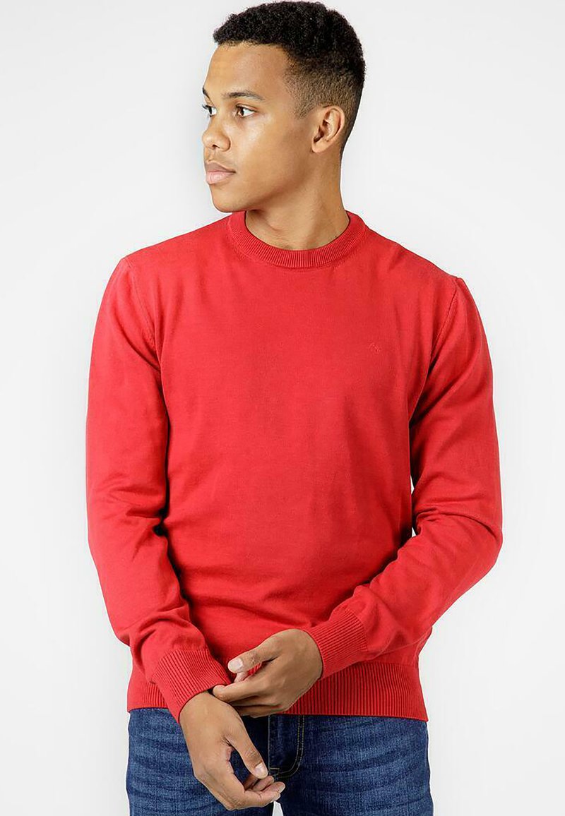 Czerwony bawełniany sweter z okrągłym dekoltem i długimi rękawami, z prążkowanymi mankietami i paskiem w talii, w zestawie z niebieskimi dżinsami. Prosty fason, bez wzorów ani grafik.