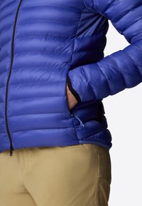 Giacca puffa blu con cuciture orizzontali e zip nera, dotata di tasca laterale e polsini elastici. Indossata sopra pantaloni di colore chiaro.