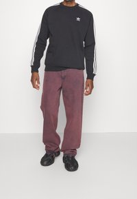 Man i svart Adidas-tröja med vita ränder på ärmarna, urtvättade lila lösa jeans och svarta sportskor står mot vit bakgrund.