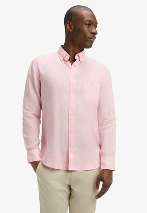 Homme portant une chemise à manches longues rose pâle avec poche poitrine, pantalon beige, debout avec une main dans la poche, regardant vers la droite.