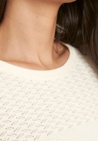 Bílý pletený svetr s texturovaným vzorem cik-cak a širokým kulatým výstřihem, vyrobený z měkkého, lehkého materiálu.