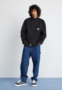 Black button-up shirt, relaxed fit with a chest pocket. Paired with loose blue denim jeans and navy sneakers. Simple and functional design.