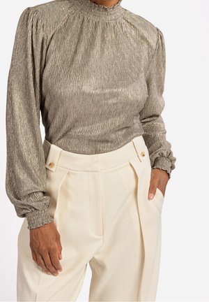 Kobieta w metalicznej bluzce z długim rękawem ze ściągaczami na mankietach oraz kremowych, wysokiegodzieżowych spodniach z guzikowymi detalami, z rękami spoczywającymi po bokach.