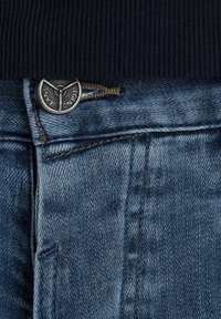 Gros plan sur la ceinture d'un jean en denim bleu avec un bouton métallique gravé "Legend Jeans" et des détails de coutures visibles.