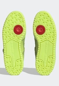Jasně žlutozelené podrážky tenisek Adidas s kruhovými dezény a centrálními červenými spirálovými gripy, zobrazené zespodu na světlém pozadí.