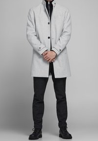 Abrigo de lana gris claro con cuello alto, cierre de botones en la parte delantera y bolsillos laterales, usado sobre un suéter negro y unos pantalones de cuadros oscuros. Botas negras.