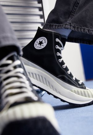 Baskets hautes Converse All Star noires avec semelle blanche épaisse, lacets blancs et écusson visible, portées avec un pantalon gris sur un sol bleu.