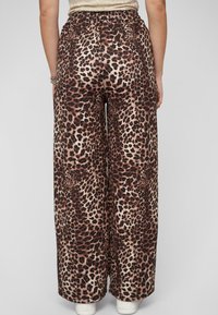 Wijd-uitlopende broek in een stoffen met luipaardprint, met bruin en zwarte vlekken op een beige achtergrond. Elastische tailleband en witte schoenen zichtbaar.