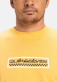 Yellow cotton t-shirt featuring a graphic logo in black and white with a checkered border. Round neckline, short sleeves.