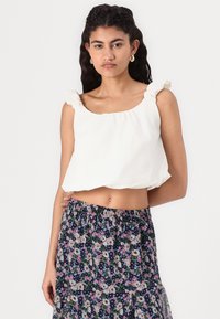 Top blanc court avec des bretelles volantes, associé à une jupe bleu marine à motifs floraux avec des accents roses et verts.