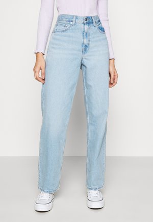 Žena nosící světle modré džíny s vysokým pasem a přímo střiženými nohavicemi, bílé tenisky a levandulové žebrované tričko s dlouhým rukávem, stojící na jednobarevném pozadí.