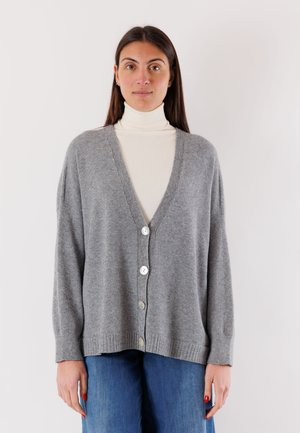 Donna che indossa un dolcevita bianco e un cardigan grigio largo con bottoni, in piedi con le mani rilassate lungo i fianchi davanti a uno sfondo semplice.