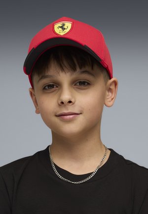 Boy with short brown hair wearing red Ferrari cap, black shirt, and silver chain necklace against a neutral gray background.