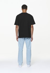 Juodas medvilninis T-shirt su trumpomis rankovėmis, apvaliu kaklu, rodomas iš nugaros. Suderintas su šviesiai mėlynomis tiesiomis kelnėmis ir baltomis sportbačiais.