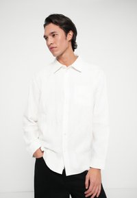 Νεαρός άνδρας με σκούρα μαλλιά που φοράει λευκό πουκάμισο με υφή και μαύρο παντελόνι, στέκεται με το ένα χέρι στην τσέπη, κοιτάζοντας προς τα αριστερά.