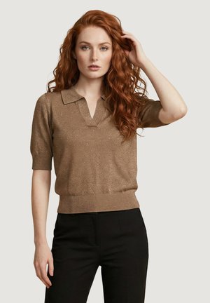 Kobieta o długich, kręconych rudych włosach, ubrana w brązową, krótkim rękawem, dzianinową bluzkę i czarne spodnie, dotyka włosów jedną ręką.