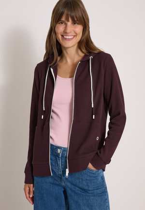 Femme souriante portant un sweat à capuche violet foncé zippé par-dessus un haut rose et un jean bleu, la main droite dans la poche.