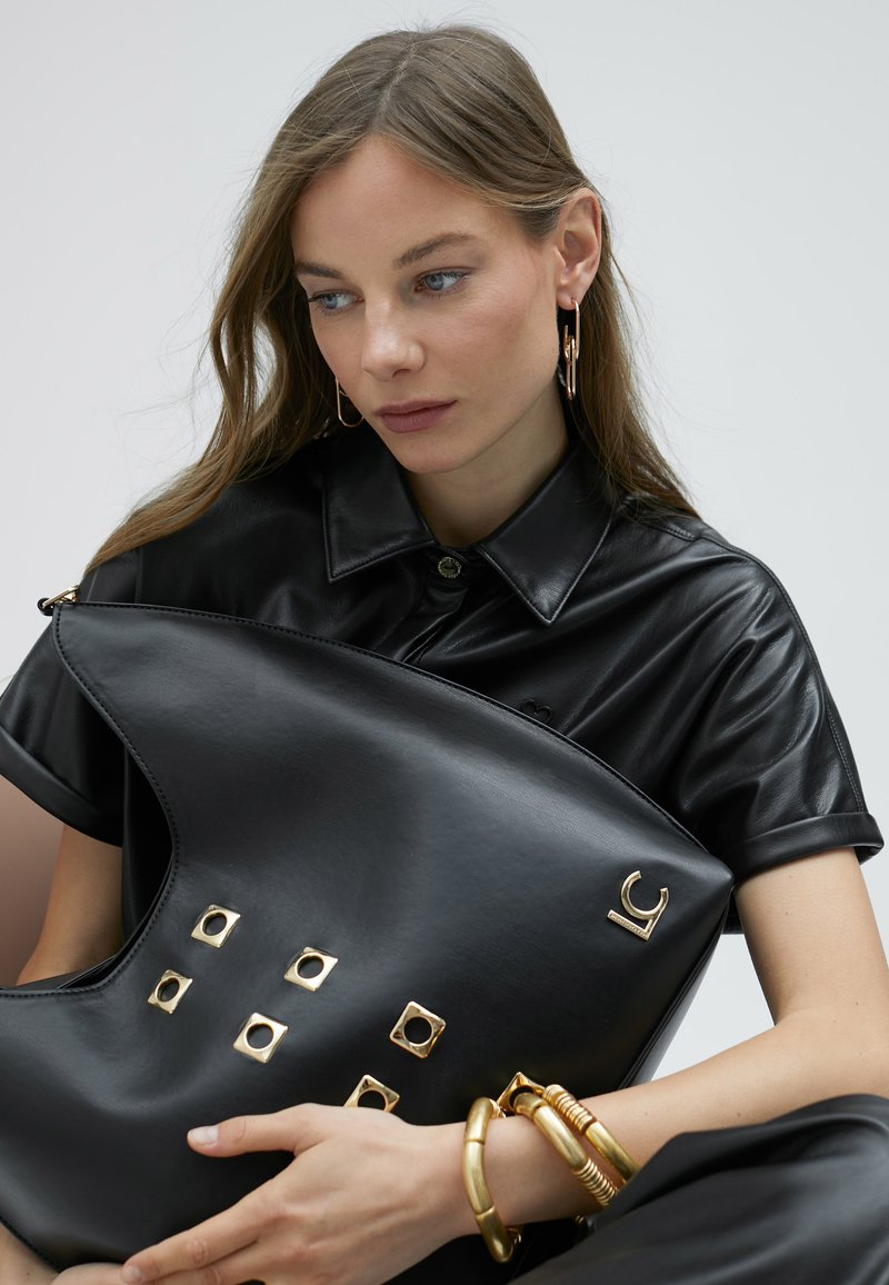 Černá kožená kabelka se zlatými čtvercovými hřebíky a logem. Hladký, zakřivený tvar s matným povrchem, doplněný zlatými náramky na zápěstí.