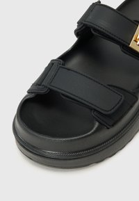 Black sandal featuring a closed toe design, two adjustable straps, smooth texture, and a slight platform sole. Accented with gold hardware.