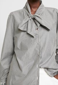 Camicia a righe bianca e nera, con un grande colletto annodato e tessuto morbido, dotata di bottoni visibili sulla parte anteriore.