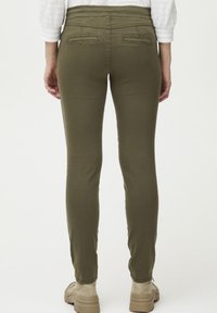 Pantalon vert olive avec une coupe slim, fabriqué dans un tissu flexible. Dispose de deux poches arrière et d'une texture lisse. Associé à des chaussures beiges.