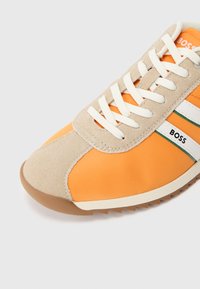 Oranger Sportschuh mit beigem Veloursleder- und Nylon-Obermaterial, weißer Schnürung, grünen und weißen Streifen sowie Gummisohle mit Profil.