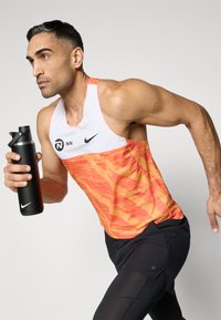 Nike tank top, millel on valge ülemine osa apelsiniga lainelise mustriga, kombineeritud mustade lühikeste pükstega. Mudel hoiab käes musta isoleeritud veepudelit.