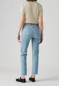 Mujer de pie de espaldas vistiendo jeans de tiro alto azul claro, top de manga corta beige y zapatos planos negros, contra un fondo liso.