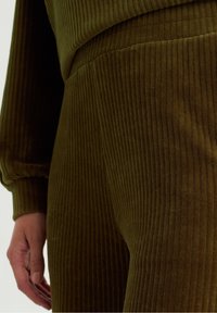 Olivgrüner ribbeliger Stoffset mit langen Ärmeln und elastischen Bündchen, das die Textur und Konturen des Kleidungsstücks sowie die Hand des Modells zeigt.