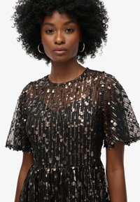 Vestido negro con lentejuelas, de mangas cortas y transparentes. Presenta un escote redondo y un patrón texturizado de lentejuelas metálicas sobre un tejido transparente.