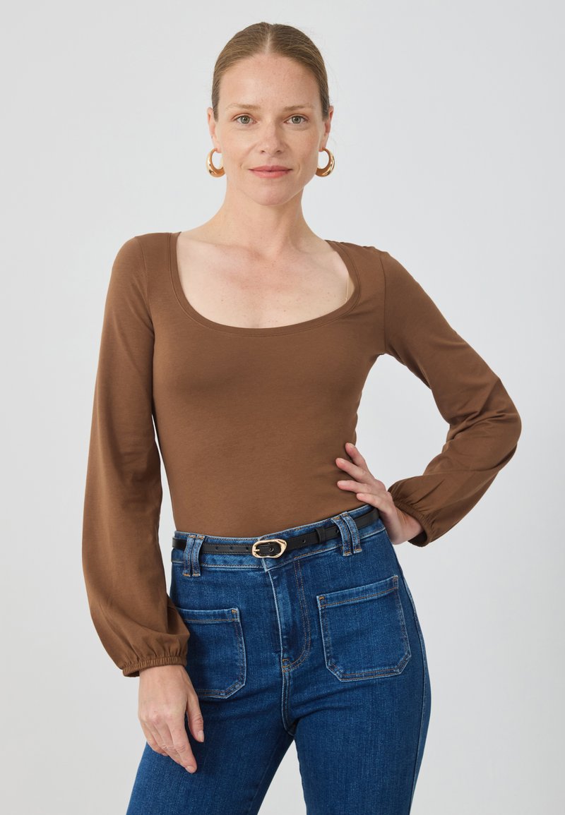 Brauner, langärmeliger Body mit quadratisch ausgeschnittenem Dekolleté, kombiniert mit hochgeschnittenen blauen Jeans aus Denim, die einen schwarzen Gürtel mit runder Schnalle tragen.